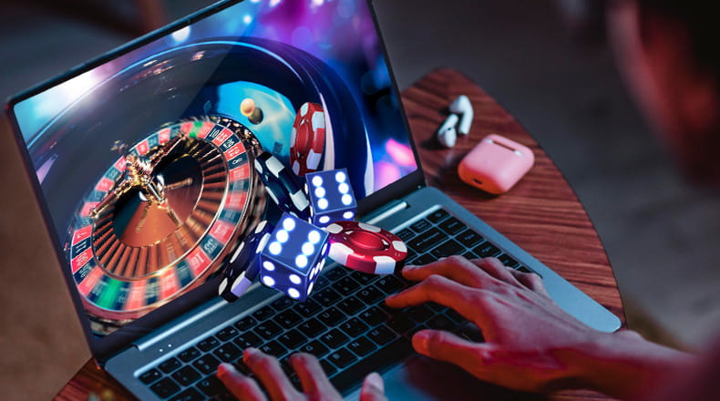 A person using a laptop to play online, with a roulette wheel, dice, and poker chips coming out of the screen.
